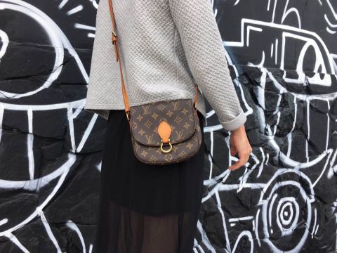 Eine Person, die einen strukturierten grauen Pullover und einen schwarzen Rock trägt, trägt eine braune Designerhandtasche mit einem Monogrammmuster. Im Hintergrund ist eine schwarze Wand mit weißen abstrakten Graffitis zu sehen.
