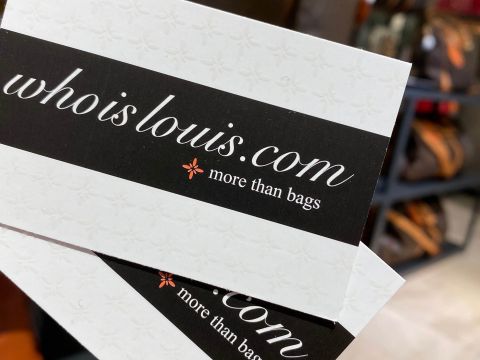 Nahaufnahme von zwei Visitenkarten mit dem Text „whoislouis.com“ und dem Satz „more than bags“ auf schwarz-weißem Hintergrund. Die Karten weisen ein dezentes Blumenmuster auf. Im Hintergrund sind Regale mit verschiedenen Taschen zu sehen.