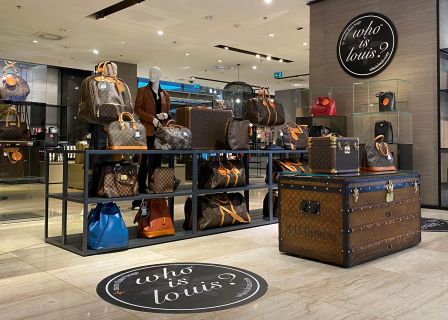 Ein Luxus-Ladendisplay, das verschiedene Ledertaschen und Koffer mit einem unverwechselbaren Monogrammmuster präsentiert. Der elegante Innenraum verfügt über Schaufensterpuppen und Regale mit Produkten. Im Vordergrund ist ein großer Koffer auffällig dargestellt.