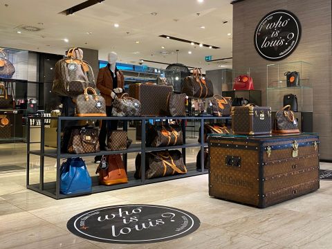 Ein Luxus-Ladendisplay, das verschiedene Ledertaschen und Koffer mit einem unverwechselbaren Monogrammmuster präsentiert. Der elegante Innenraum verfügt über Schaufensterpuppen und Regale mit Produkten. Im Vordergrund ist ein großer Koffer auffällig dargestellt.