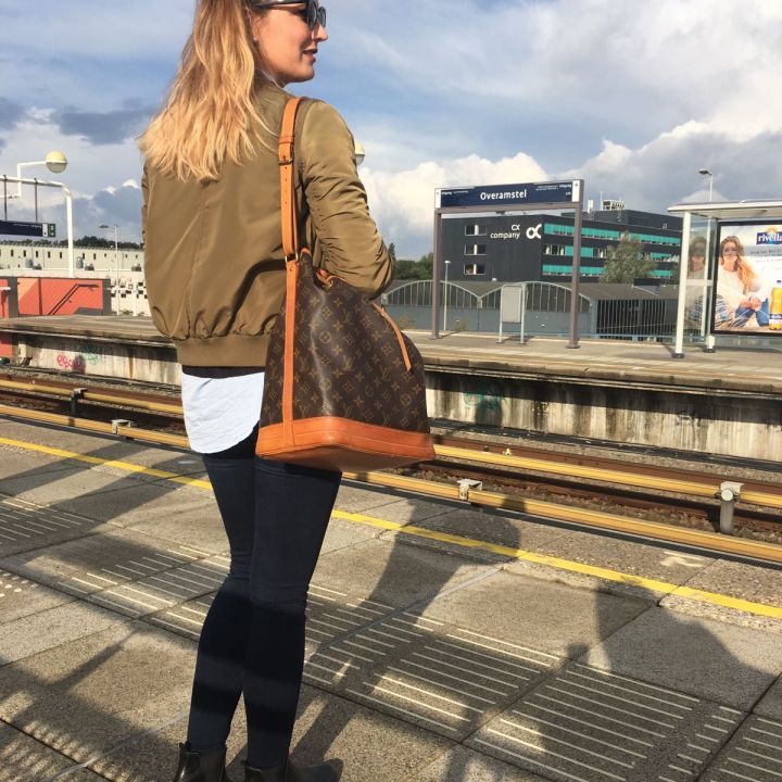 Eine Frau steht auf einem Bahnsteig und blickt in die Ferne. Sie trägt eine Sonnenbrille, eine grüne Jacke, Jeans und schwarze Stiefel und hat eine braune Tasche über der Schulter. Der Himmel ist teilweise bewölkt und im Hintergrund ist ein Gebäude zu sehen.