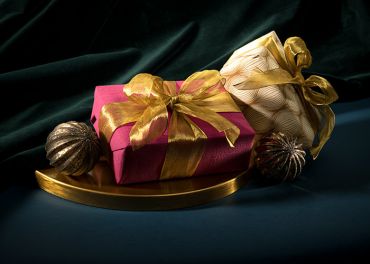 Zwei elegant verpackte Geschenke, eines in Rot und eines in Gold, mit goldenen Bändern zusammengebunden, liegen auf einer geschwungenen goldenen Oberfläche. Sie werden von zwei dekorativen, strukturierten Bronzekugeln begleitet. Der Hintergrund ist dunkelgrüner Samt, der das luxuriöse Gefühl verstärkt.