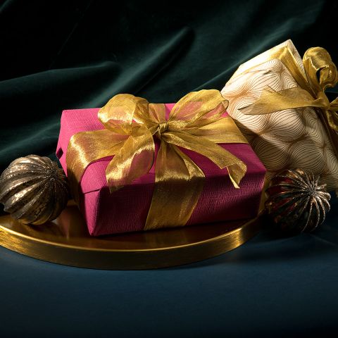Zwei elegant verpackte Geschenke mit goldenen Bändern liegen auf einem goldenen Tablett vor einem dunkelgrünen Stoffhintergrund. Das linke Geschenk ist in rotes Papier eingewickelt, das rechte in cremefarbenes Papier mit Wellenmuster. Zwei strukturierte Kugeln flankieren sie.