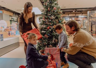 Eine Familie schmückt drinnen einen Weihnachtsbaum. Eine Frau hält ein großes rotes Geschenk, ein Mann hockt daneben und zwei Kinder schauen aufgeregt auf die verpackten Geschenke unter dem Baum. Festliche Dekorationen und warmes Licht sorgen für eine fröhliche Weihnachtsstimmung.