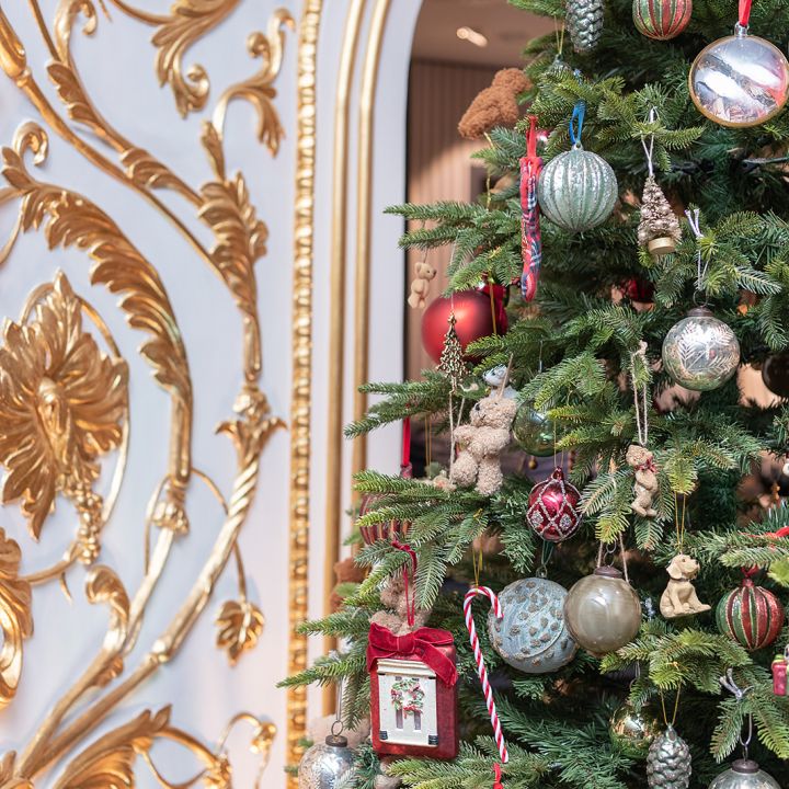 Ein festlicher Weihnachtsbaum, geschmückt mit verschiedenen bunten Ornamenten, darunter Kugeln, eine Zuckerstange und ein kleiner Bilderrahmen. Der Hintergrund besteht aus einer verzierten, goldfarbenen Wand mit dekorativen Mustern, die eine luxuriöse Feiertagsatmosphäre schaffen.