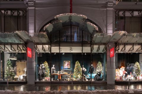 Ein festliches Schaufenster mit Weihnachtsdekorationen, darunter funkelnde Lichter, Weihnachtsbäume und Schaufensterpuppen in Winterkleidung. Das Fenster wird von einer gewölbten Struktur eingerahmt und der Boden draußen ist leicht mit Schnee bedeckt.