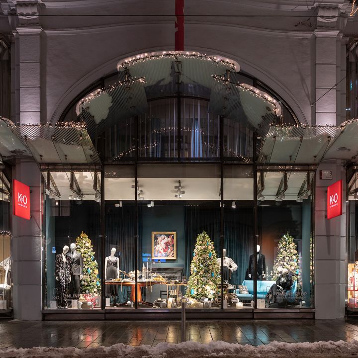 Ein festliches Schaufenster mit Weihnachtsdekorationen, darunter funkelnde Lichter, Weihnachtsbäume und Schaufensterpuppen in Winterkleidung. Das Fenster wird von einer gewölbten Struktur eingerahmt und der Boden draußen ist leicht mit Schnee bedeckt.