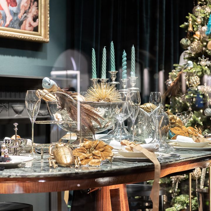 Ein üppig dekorierter Esstisch für einen festlichen Anlass mit grünen Kerzen, goldenen Akzenten, Kristallglas und einem luxuriösen Weihnachtsbaum im Hintergrund. Das Ambiente ist elegant mit einer dunklen Wand und einem gerahmten Gemälde.