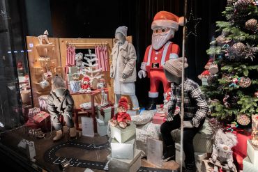 In einem festlichen Schaufenster sind Schaufensterpuppen in Winterkleidung zu sehen, umgeben von Geschenkkartons, Spielzeug und einer großen Weihnachtsmannfigur. Auf der rechten Seite steht ein geschmückter Weihnachtsbaum, der das Weihnachtsthema noch verstärkt.