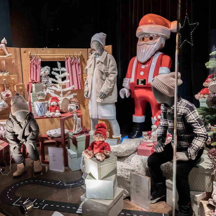 In einem festlichen Schaufenster sind Schaufensterpuppen in Winterkleidung zu sehen, umgeben von Geschenkkartons, Spielzeug und einer großen Weihnachtsmannfigur. Auf der rechten Seite steht ein geschmückter Weihnachtsbaum, der das Weihnachtsthema noch verstärkt.