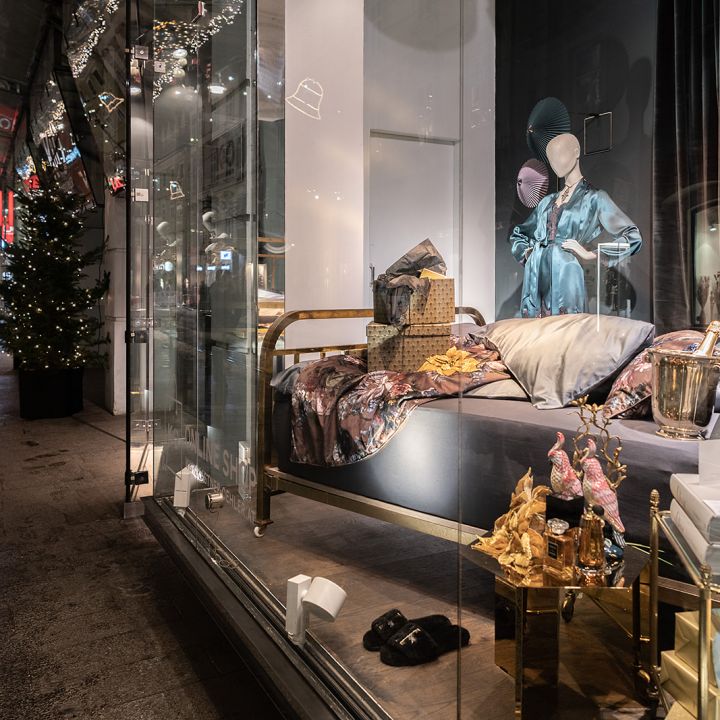Ein Schaufenster mit einem elegant gemusterten Bett und verschiedenen Luxusartikeln wie Handtaschen und einem Champagnerkühler. Weihnachtsbeleuchtung und Dekorationen schmücken die Straße draußen und sorgen für festliche Atmosphäre.