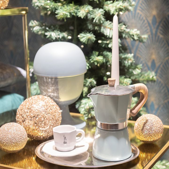 Ein festliches Arrangement besteht aus einer Espressomaschine für den Herd, einer weißen Tasse auf einer Untertasse und einer brennenden Kerze auf einem Tablett. Um sie herum sind glitzernde goldene Ornamente und ein kleiner Weihnachtsbaum vor einer dekorativen Wand aufgestellt.