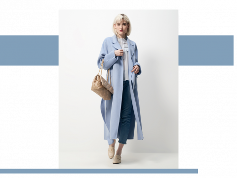 Eine Person mit kurzen hellen Haaren trägt einen langen blauen Mantel, Jeans und Halbschuhe. Sie steht vor einem weißen Hintergrund mit blauen Akzentstreifen und hält eine hellbraune gesteppte Handtasche.