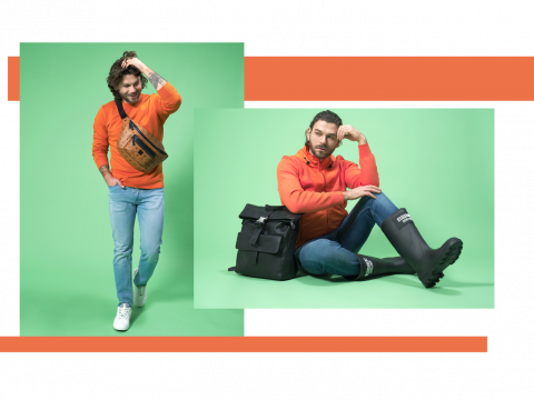 Zwei Männer in orangefarbenen Pullovern posieren vor einem grünen Hintergrund. Der erste trägt Jeans, weiße Turnschuhe und eine braune Umhängetasche. Der zweite sitzt mit einem schwarzen Rucksack da und trägt dunkle Gummistiefel. Beide stehen vor einem sich überschneidenden orangefarbenen und grünen Hintergrund.
