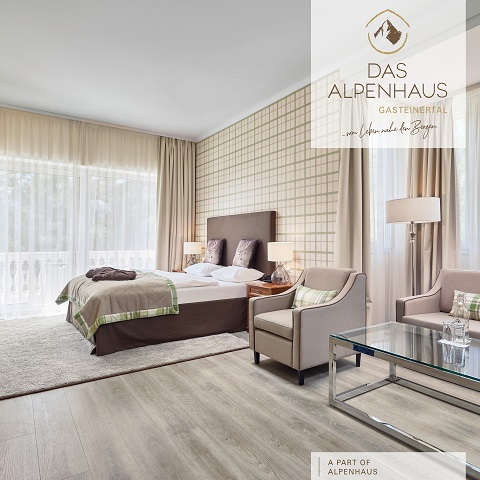 Elegantes Hotelzimmer mit Doppelbett, moderner Einrichtung und neutralen Farbtönen. Zur Ausstattung gehören ein Couchtisch mit Glasplatte, Sessel und ein großes Fenster mit transparenten Vorhängen, die den Raum mit natürlichem Licht durchfluten. Text lautet „Das Alpenhaus Gastenerial“.
