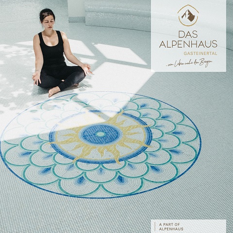 Eine Frau sitzt im Schneidersitz meditierend auf einem gefliesten Boden mit einem bunten Mandala-Muster. Sonnenlicht fällt durch ein Fenster. In der oberen rechten Ecke sind die Worte „Das Alpenhaus Gasteinertal“ zu sehen.