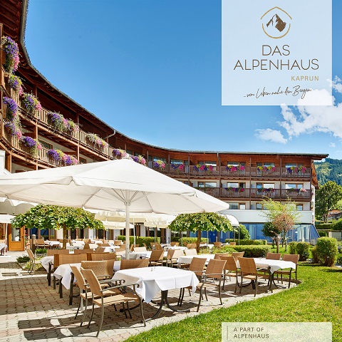 Gastgarten im Alpenhaus Kaprun mit Tischen mit weißen Tischdecken und großen Sonnenschirmen. Im Hintergrund ist das Hotelgebäude mit blumengeschmückten Holzbalkonen unter einem klaren blauen Himmel zu sehen.