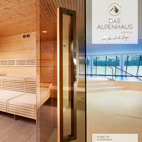 Innenansicht eines ruhigen Saunaraums mit Holzbänken und warmer Beleuchtung. Eine Glastür öffnet sich und gibt den Blick auf einen geräumigen Bereich frei. Draußen blicken große Fenster auf eine üppige grüne Landschaft. Das Branding für „Das Alpenhaus“ ist sichtbar.