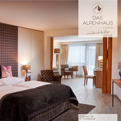 Ein gemütliches Hotelzimmer mit einem großen Bett, Holzakzenten und einer Sitzecke mit zwei Stühlen und einem kleinen Tisch. Sanfte Beleuchtung verstärkt die warme, einladende Atmosphäre. Ein Fenster mit transparenten Vorhängen lässt natürliches Licht herein.