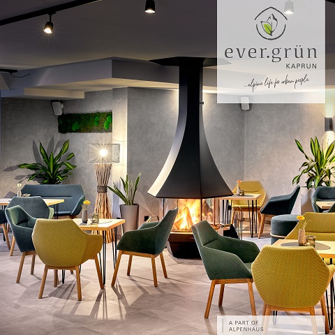 Ein moderner Loungebereich mit grünen und gelben Stühlen um kleine Holztische. In der Mitte steht ein schwarzer, kegelförmiger Kamin. Der Raum ist von Pflanzen umgeben. Das Bild zeigt das Logo von „ever.grün Kaprun“ mit dem Slogan „Alpines Leben für Stadtmenschen“.