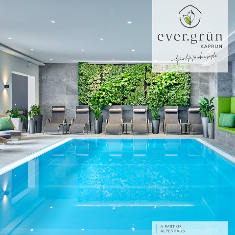Innenpoolbereich mit Liegestühlen und einem vertikalen Garten an der Wand. Die Umgebung ist modern und hell, mit Topfpflanzen und eleganten Designelementen. Der Markentext lautet „ever.grün Kaprun“ und „ein Teil von Alpenhaus“.
