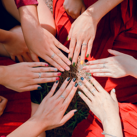 Ein Kreis unterschiedlicher Hände zeigt farbenfrohe Maniküren und Ringe über Gras. Jede Hand zeigt einzigartige Nagelkunst, darunter schwarze, rote und grüne Designs, und trägt verschiedene silberne Ringe. Die Hände liegen auf einem roten Stoffhintergrund.