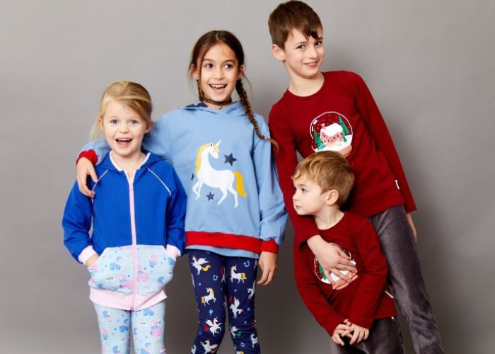 Vier Kinder stehen zusammen vor einem grauen Hintergrund. Sie tragen bunte Pyjamas, einer mit Einhorn-Motiv, der andere mit festlichem Aufdruck. Sie wirken fröhlich und entspannt.