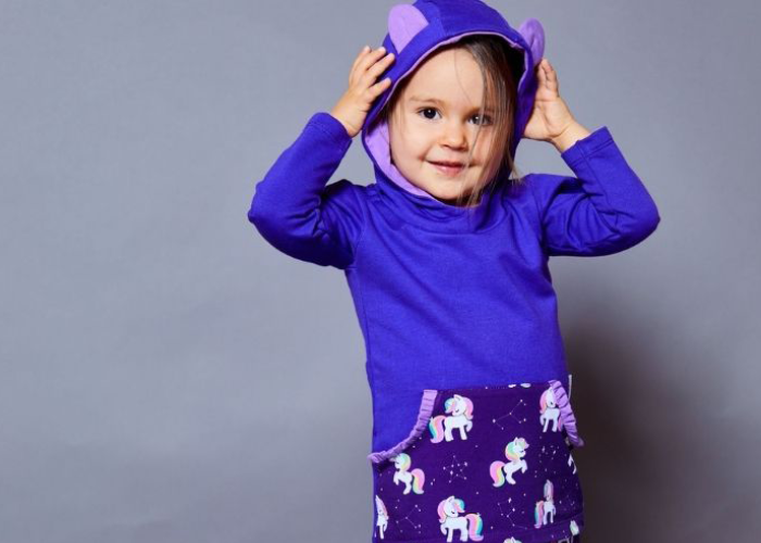 Ein Kind in einem lila Kapuzenpullover mit Bärenohren und Einhornmuster auf der Tasche lächelt vor einem schlichten grauen Hintergrund.