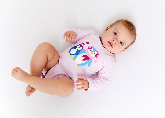 Ein Baby in einem rosa Strampler mit bunten Pinguinmotiven liegt auf dem Rücken vor einem weißen Hintergrund. Das Baby hat kurzes, hellbraunes Haar und schaut nach oben.