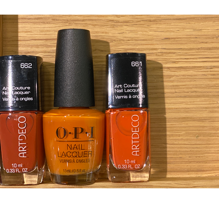 Auf einer Holzfläche sind drei Nagellackfläschchen ausgestellt. Die linke und rechte Flasche sind von Artdeco, Farbtöne 662 (dunkelrötlich) und 661 (hellorange), während die mittlere Flasche ein OPI in einem leuchtenden Orange ist.