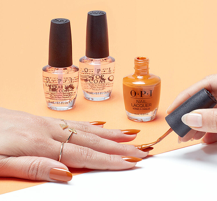 Eine Person trägt orangefarbenen Nagellack auf ihre Fingernägel auf. Im Hintergrund stehen zwei OPI-Nagellackflaschen auf einer orangefarbenen Oberfläche. Die Hand trägt einen eleganten goldenen Ring und die Nägel sind sorgfältig manikürt.
