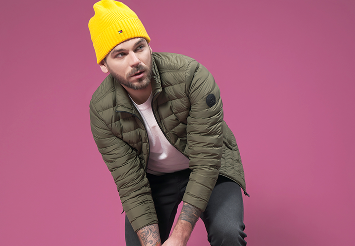 Eine Person mit einer gelben Beanie, einer olivgrünen Pufferjacke, einem weißen Hemd und dunklen Hosen posiert vor einem rosa Hintergrund. Sie lehnt sich leicht nach vorne und hat einen konzentrierten Gesichtsausdruck.