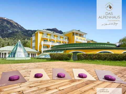 Outdoor-Yogabereich mit Matten und Kissen auf einem Holzdeck vor einem gelben Hotelgebäude mit Bergblick. Im Hintergrund sind grüne Dächer und eine gläserne Pyramidenstruktur zu sehen.
