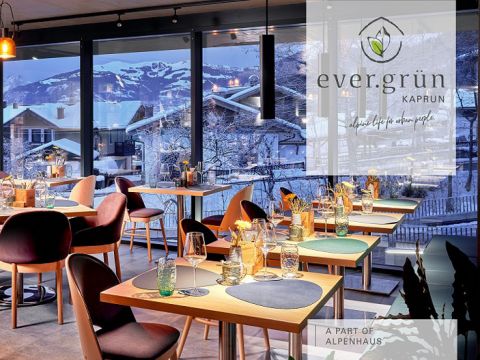 Ein moderner Restaurant-Essbereich mit Holztischen und -stühlen, gedeckt mit Gläsern und Tischsets, bietet durch große Fenster einen Blick auf die schneebedeckten Berge. Über dem Slogan „Indoor-Leben der Stadtmenschen“ steht das Logo „ever.grün Kaprun“.