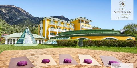 Auf einem Holzdeck mit Blick auf ein gelbes Hotelgebäude, das von üppigen grünen Bergen umgeben ist, sind Yogamatten und -kissen für draußen ausgelegt. Das Gebäude hat eine pyramidenförmige Glasstruktur und ein rundes grünes Dach. Der Himmel ist klar und sonnig.