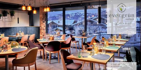 Gemütliches Restaurant-Interieur mit Holztischen zum Essen und Blick durch große Fenster auf ein verschneites Bergdorf. Warme Pendelleuchten hängen von der Decke und schaffen eine einladende Atmosphäre. In der Ecke ist das Logo „ever.grün Kaprun“ zu sehen.