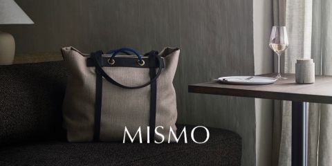 Eine schicke beigefarbene Einkaufstasche mit schwarzen Griffen liegt auf einem dunklen Sofa. Daneben steht ein Holztisch mit einem Weinglas und einem Teller. Das Ambiente wirkt elegant und minimalistisch, das Wort „Mismo“ ist prominent platziert.