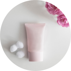 Eine pastellrosa Tube Kosmetikcreme auf einer weißen Oberfläche, begleitet von drei kleinen weißen Wattebällchen. In der oberen rechten Ecke sind teilweise rosa Blumen zu sehen. Die Komposition ist minimalistisch und sauber.