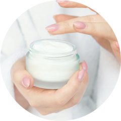 Eine Person mit sorgfältig manikürten Nägeln hält ein offenes Gefäß mit weißer Creme und ist bereit, sie mit dem Finger aufzutragen. Der Hintergrund ist weich und verschwommen und suggeriert ein beruhigendes Selbstpflegeritual.