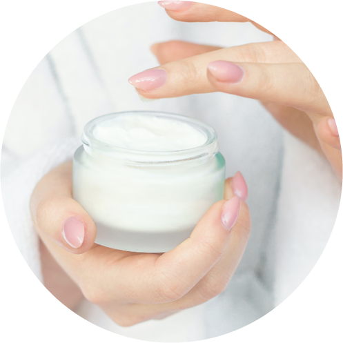 Eine Person mit sorgfältig manikürten Nägeln hält ein offenes Gefäß mit weißer Creme und ist bereit, sie mit dem Finger aufzutragen. Der Hintergrund ist weich und verschwommen und suggeriert ein beruhigendes Selbstpflegeritual.