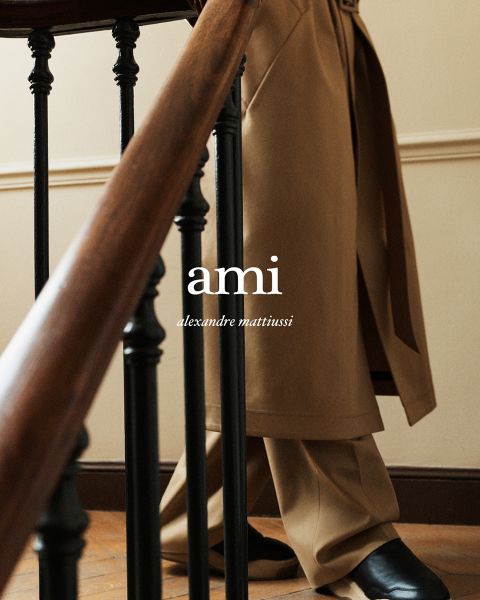 Eine Person in beigem Mantel und Hose steht hinter einem hölzernen Treppengeländer. Der Hintergrund ist eine helle Wand. Der Text auf dem Bild lautet „ami alexandre mattiussi“. Der Fokus liegt auf der Kleidung und der Treppe.