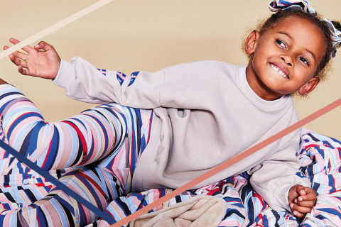 Ein lächelndes kleines Kind liegt auf einer bunten, gestreiften Decke. Es trägt ein helles Sweatshirt und gestreifte Leggings, die zur Decke passen. Der Hintergrund ist neutral und betont die leuchtenden Farben.