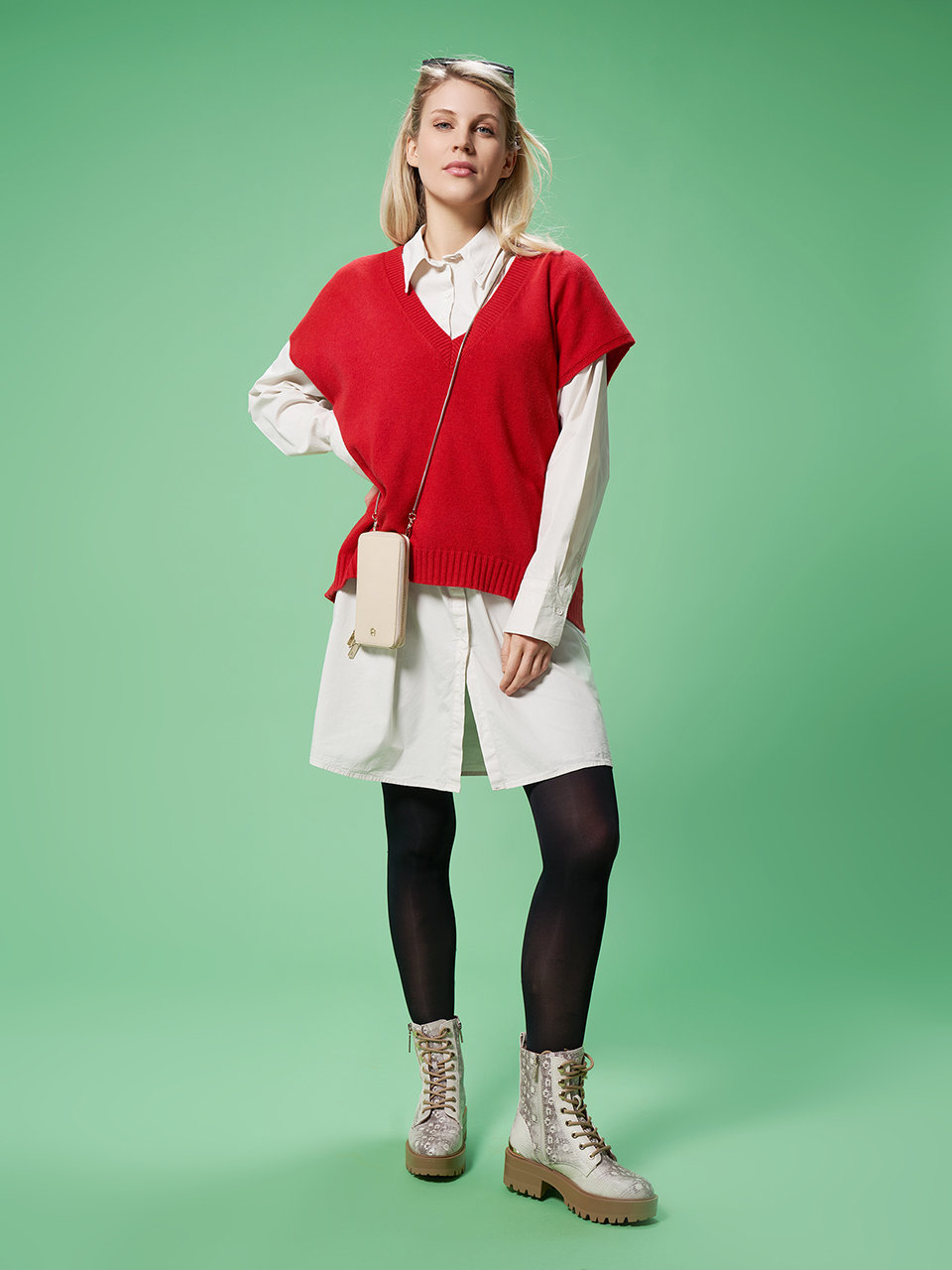 Eine Person steht vor einem grünen Hintergrund und trägt einen roten Pullover über einem weißen Hemdblusenkleid, schwarze Leggings und beige Stiefel. Sie trägt eine kleine Umhängetasche und eine Sonnenbrille auf dem Kopf.