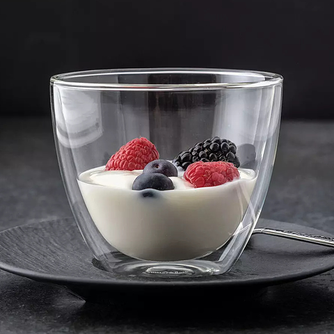 Ein doppelwandiger Glasbecher ist mit cremigem Joghurt gefüllt und mit frischen Himbeeren, Brombeeren und Blaubeeren garniert. Der Becher steht auf einer schwarzen Untertasse mit einem Löffel daneben, vor einem dunklen Hintergrund.