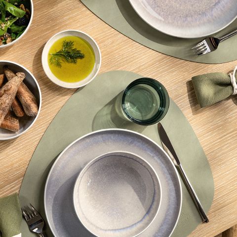 Draufsicht auf einen Esstisch mit grauen Tellern, grünen Tischsets, einer Schüssel Grissini, einer Schüssel grüner Dillsauce und einem kleinen grün getönten Glas. Servietten sind ordentlich gerollt und neben den Tellern platziert.