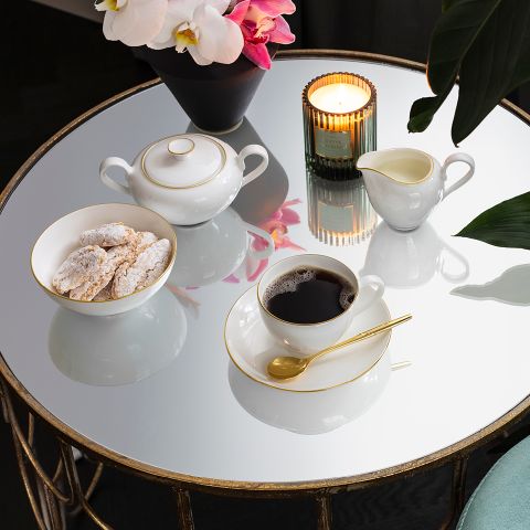 Auf einem runden, verspiegelten Tisch stehen eine brennende Kerze, eine Tasse Kaffee mit Untertasse und goldenem Löffel, eine Schüssel mit Keksen mit Puderzucker, eine Zuckerdose, ein Sahnekännchen und eine Vase mit rosa und weißen Orchideen.