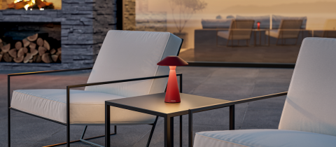 Eine gemütliche Außenterrasse mit zwei modernen weißen Sesseln und einer roten Lampe auf einem kleinen schwarzen Tisch, vor der Kulisse eines Sonnenuntergangs und einem Kamin mit gestapelten Holzscheiten. Eine Glastür gibt den Blick auf weitere Sitzgelegenheiten im Inneren frei.