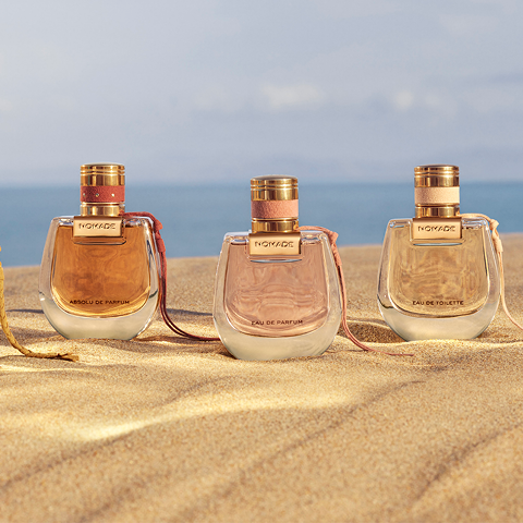 Drei Parfümflaschen mit der Aufschrift „Nomade“ stehen auf goldenem Sand. Die Flaschen haben ein geschwungenes Design mit goldenen Verschlüssen und enthalten jeweils einen anderen Duft. Der Hintergrund ist ein klarer blauer Himmel und ein verschwommener Ozean, was ein Gefühl von Luxus und Ruhe vermittelt.