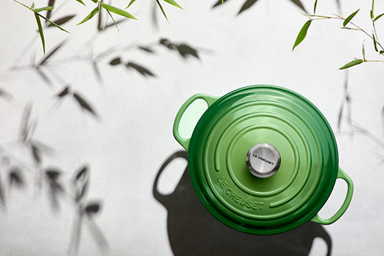 Ein grüner Le Creuset-Topf mit silbernem Knauf auf einer hellen Oberfläche, die Blätterschatten wirft.