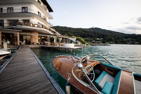 Ein hölzernes Motorboot liegt an einem Steg am Seeufer in der Nähe eines luxuriösen Hotels. Das Hotel verfügt über einen Balkon und Sitzgelegenheiten im Freien mit Blick auf die grünen Hügel und das ruhige Wasser unter einem teilweise bewölkten Himmel.